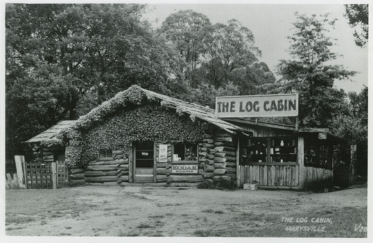 Shows The Log Cabin in Marysville in Victoria. Shows the front view of The Log Cabin with a vine growing across the front of the facade. To the right of the building is a large sign and there are advertising signs on the front of the cabin.