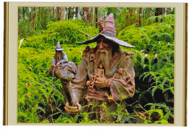Shows a terracotta sculpture of a bearded wizard wearing a tall hat and carrying a staff that has a smaller wizard sitting on top of it. There are several small figures sitting on and around the large wizard. The wizard also has a frog sitting on his left shoulder