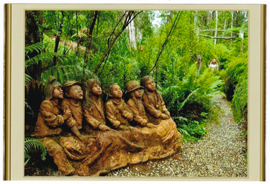 Shows a terracotta sculpture of a group of six children, three boys and three girls, all seated on a long seat. They are dressed in old fashioned clothing with three of them wearing different hats. They are all looking up and one of the boys has stuck his tongue out