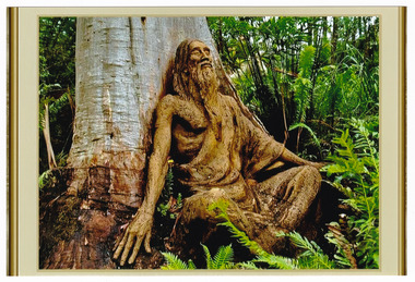 Shows a terracotta sculpture of a guru type bearded man sitting cross-legged against the trunk of a large tree. The man has long hair and is wearing a type of robe. His arms are resting on the ground next to his knees. He appears to be meditating.