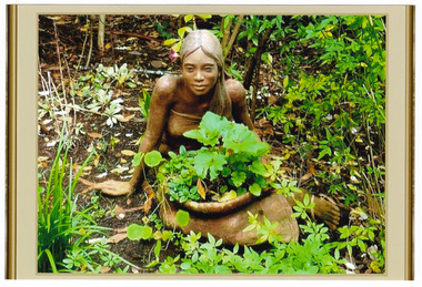 Shows a terracotta sculpture of a woman sitting on the ground behind a large sack shaped plant pot. The woman has long hair and is wearing a dress.