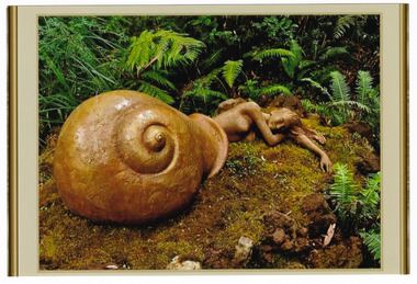 Shows a terracotta sculpture of a naked women lying with her lower body and legs under a large smail shell. The lady is lying prone with her head resting on her arms.