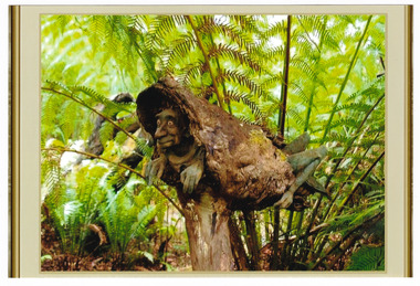 Shows a terracotta sculpture of a grasshopper like creature with the head of a human. The creature is encased in a piece of an old log with its head protruding out of one end and its legs and tail out the other.