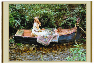 Shows a terracotta sculpture of a woman sitting in a small wooden boat. She is dressed in a long dress that is belted at her hips and she is wearing a head band in her long hair. One hand is resting in her lap and the other is holding what appears to be a fishing line. Her feet are resting on a large woven floral rug that is folded in the bottom of the boat. In the bow of the boat is a small lantern. The boat is tied up and is lying on the bank next to a small creek. 