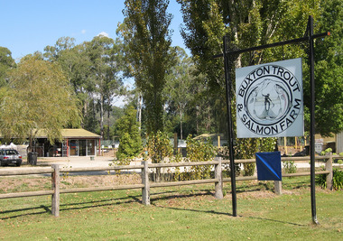 Shows the sign out the front of the Buxton Trout & Salmon Farm. Shows a building with a carpark surrounded by a wooden fence.
