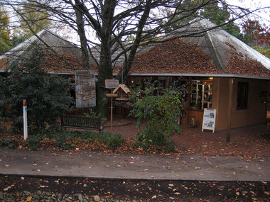 Shows the front facade of Country Touch in Marysville in Victoria. Shows a sign made of corregated iron with wooden signs with the goods and services available at the shop. Shows also two large wooden bird feeders. There is also a green wooden bench seat in the front yard of the shop.