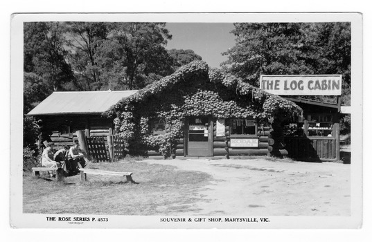 Shows the front of The Log Cabin in Marysville in Victoria. In the left of the photograph are a women and two men sitting on one of three wooden bench seats.