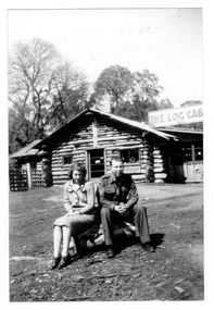 Shows the front facade of The Log Cabin in Marysville in Victoria. There is a man and a woman sitting on a large rock in front of the building. The man is in battledress uniform.