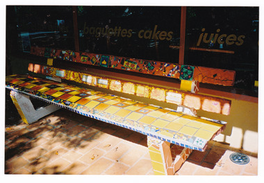 Shows a wooden bench seat covered in mosaic tiles.