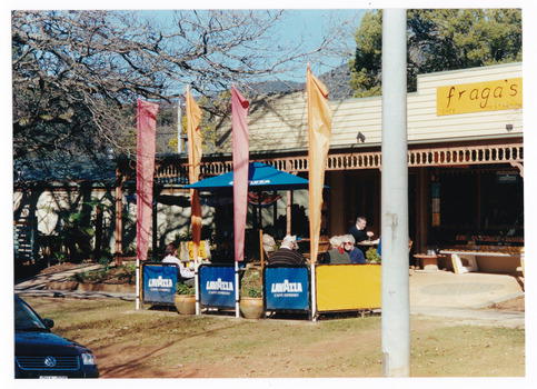 Shows the front facade of Fraga's cafe restaurant in Marysville in Victoria. Shows a bench seat out the front with further seating with flags on the outside of the footpath. 
