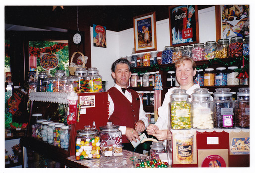 Shows Uncle Fred & Aunty Val's Old Style Lolly Shop in Marysville in Victoria. Shows Frank and Val Pryor standing behind the counter in the Lolly Shop. Shows jars and jars of lollies both on the counter and on shelving behind the counter. On the wall behind the counter there are advertising posters advertising chocolate and coke-a-cola.
