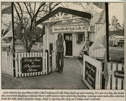 Shows the owner of the Marysville Old Style Lolly Shop standing outside the shop's temporary premises after the 2009 Black Saturday bushfires destroyed the original building. Shows the fence and gateway arbor along with the signage that survived the fires.