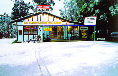 Shows the front facade of Narbethong Ski Hire in Victoria during winter. Shows a building with signs at the front advertising goods and services available at the shop. The building is surrounded by snow.