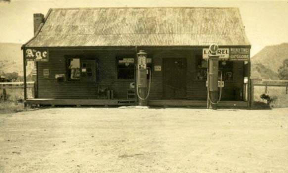 Shows a black and white photograph of the Taggerty General Store in Victoria. Shows a weatherboard building with a corregated iron roof and a brick chimney. Shows two petrol bowsers standing out the front of the building. In both corners of the verandah are advertising signs for the Age newspaper and the Weekly Times.