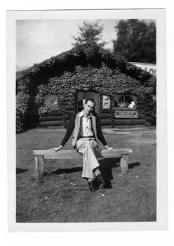 Shows a man sitting on a wooden bench seat outside the front of The Log Cabin in Marysville in Victoria. On the front facade of the building is an advertising sign for Kodak Supplies and another for Red Capstan cigarettes.