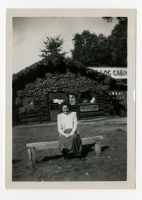 Shows a lady sitting on a wooden bench seat outside the front of The Log Cabin in Marysville in Victoria. On the front facade of the building is an advertising sign for Kodak Supplies and another for Red Capstan cigarettes.