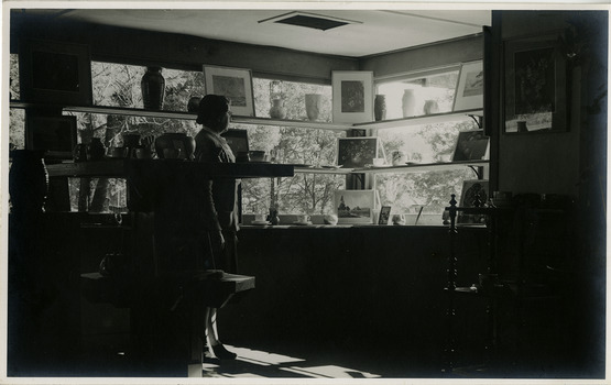 Shows a view inside The Log Cabin in Marysville in Victoria. There are shelves with various items placed on them including vases and pictures. There is a lady standing looking out at the view from the shop.