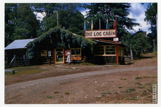 Shows The Log Cabin in Marysville in Victoria. Shows a lady standing in front of the building. Advertising signs are attached to the building for Kodak and Better Tobacco Capstans. The front verandah roofing in covered in a green leafy vine.