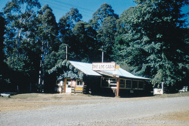 Shows The Log Cabin in Marysville in Victoria. Shows advertising signs attached to the front of the building. Shows the front verandah roof covered in a green leafy vine.