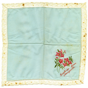 Shows a blue handkerchief with white lace surrounding the edges. In one corner there is two hand painted stems of pink lily-like flowers with the words "Greetings from Marysville" hand painted in red letters below.