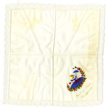 Shows a white handkerchief with a white lace edging. In one corner is a hand painted lady wearing a blue and red dress and a yellow hat. She is also carrying a blue umbrella. There is a wreath of red flowers with green and yellow leaves surrounding her. Below the picture are the words "Greetings from Marysville" in faded writing.