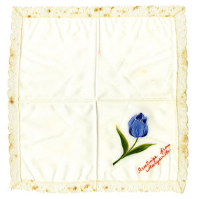 Shows a white handkerchief surrounded by white lace. In one corner is a hand painted blue tulip. Below the tulip are the words "Greetings from Marysville" hand painted in red paint.