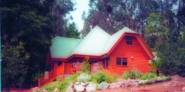 Shows one of the cottages at Anastasia's Cottages in Marysville. Shows a two storey timber cottage which is surrounded on two sides by a verandah. The cottage is set within a garden and backs onto a forest.