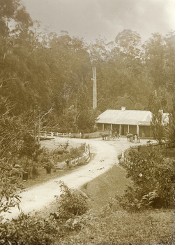 Shows a black and white photograph of the Black Spur Hotel in Narbethong. Shows a building with a verandah along the front facade. Above the front door is the sign for the hotel. In front of the hotel are two horse-drawn drays, both with people on board.