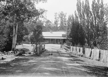 Shows a black and white photograph of J. Roches Black Spur Hotel at Narbethong. Shows a building with a verandah running along the front facade. There are two people sitting on the edge of the verandah alongside two men who are standing in front of the entry to the hotel. The photograph is taken looking down the hotel driveway at the hotel.