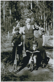 Shows the Branchflower Family in Marysville in Victoria. Shows a man standing with his arms around the shoulders of two women. Shows two young boys, one on a scooter and the other on a tricycle in front of the man and women.