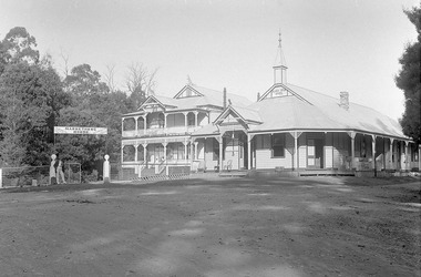 Shows a black and white photograph of the Black Spur Hotel and Narbethong House in Narbethong. The Black Spur Hotel is a weatherboard building with a verandah running along the front and left side of the building. Narbethong House is a double storey weatherboard building with verandahs on both levels. There is a set of stairs leading up to the front entrance. There is a kangaroo and a kookaburra carved from wood on the gabled peaks of the roof of Narbethong House. There is also a man standing under the sign for Narbethong House.