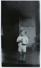 Shows a member of the Branchflower Family in Marysville in Victoria. Shows a young boy standing on the verandah of a house. He has an unknown object on his head and is holding a torch.