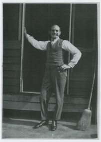 Shows a man standing on a house verandah in front of a window. He is smoking a pipe. Next to him is a straw broom.