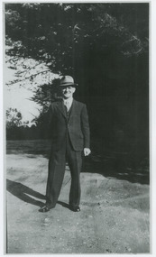Shows a man standing on a road underneath a tree. He is wearing a suit and a hat.
