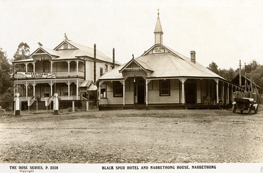 Shows a black and white photograph of the Black Spur Hotel and Narbethong House in Narbethong. The Black Spur Hotel is a weatherboard building with a verandah running along the front and left side of the building. Narbethong House is a double storey weatherboard building with verandahs on both levels. There is a set of stairs leading up to the front entrance. There is a kangaroo and a kookaburra carved from wood on the gabled peaks of the roof of Narbethong House. There is an early model car situated near the Black Spur Hotel.