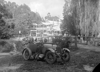 Shows a black and white photograph of Narbethong House situated next to the Black Spur Hotel in Narbethong. Narbethong House is a double storey weatherboard building with verandahs on both levels. There is a wood carved kangaroo, kookaburra and emu on the gabled peaks of the roof. In the grounds in front of Narbethong House are is a group of men and an early model car with a man standing at the rear of the car. In the foreground of the photograph is another early model car with a lady, a boy and a girl seated in the rear seat. There is another young boy seated on the running board of the car. There are two other ladies standing next to the car, one in conversation with the lady seated in the car.