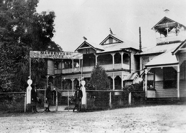 Shows a black and white photograph of Narbethong House situated next to the Black Spur Hotel in Narbethong. Narbethong House is a double storey weatherboard building with verandahs on both levels. There is a wood carved kangaroo, kookaburra and emu on the gabled peaks of the roof. There are three women standing at the entrance gateway under the Narbethong House sign. The sign advertises Luncheon, Afternoon Tea and Accommodation for Tourists.
