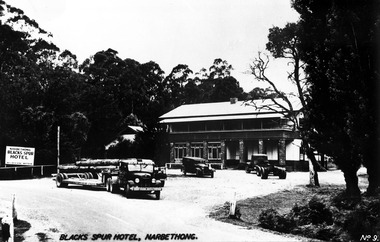 Shows a black and white photograph of the Blacks Spur Hotel in Narbethong. Shows a large double storey building with a verandah running along the front of both floors. Shows a number of vehicles standing in the front car park alongside a sign advertising the hotel.
