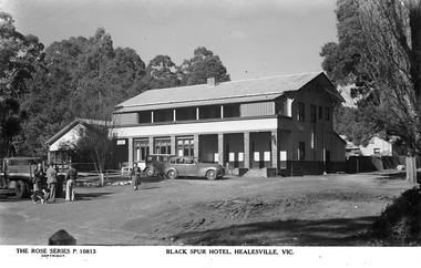 Shows a black and white postcard of the Black Spur Hotel in Narbethong. Shows a large double storey building with a verandah running along the front of both floors. Shows two cars at the entrance to the hotel with a woman walking past. Shows also a truck parked out the front with three men standing and talking with a dog standing with them.