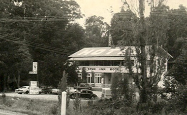 Shows a black and white photograph of the Black Spur Inn Hotel in Narbethong. Shows a large double storey building with a verandah running along the front of both floors. The word HOTEL is painted on the roof of the hotel. Shows a number of cars parked in the front carpark of the hotel.