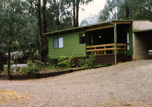 Shows one of the cottages at Blackwood Cottages in Marysville. Shows a green painted weatherboard building with a small verandah at the front. The cottage has a small garden at the front and is surrounded by large trees.