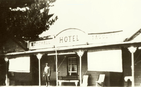 Shows the front facade of the Burchall Hotel in Taggerty. Shows a weatherboard building with a verandah running along the front. There is a man standing on the verandah. There are a variety of benches and chairs standing on the verandah. There are two pull down blinds sheltering the verandah. The sign advertising the hotel is along the roof of the verandah.