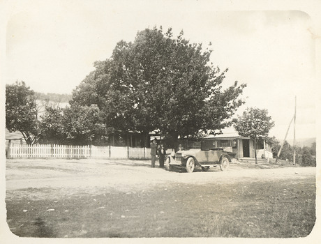 Shows two weatherboard buildings. One of the buildings is located behind a picket fence. In front of the fence is an early model car with three men standing alongside the car.