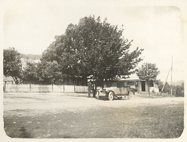 Shows two weatherboard buildings. One of the buildings is located behind a picket fence. In front of the fence is an early model car with three men standing alongside the car.