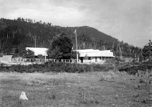 Shows a black and white photograph of Nichol's Buxton Hotel in Buxton. Shows a large building with a verandah running along the length of the building. There is a sign advertising the building along the front facade and a flag pole standing at the front fence of the building.