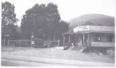 Shows a large weatherboard building with a verandah running along the length of the building. There are a set of wooden stairs leading up to the front entrance door. There is a sign above the front entrance door advertising the hotel with the name of the licensee. There are two men standing on the verandah with two other men seated on a wooden bench seat. Parked outside the front fence is a car with two men seated on a bench seat next to the car.