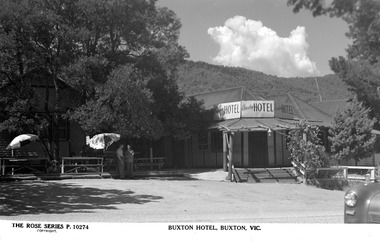 A black and white postcard of the Buxton Hotel in Buxton. Shows a large weatherboard building with a verandah running along the length of the building. There are a set of wooden stairs leading up to the front entrance door. There is a sign above the front entrance door advertising the hotel. There are two men standing in front of the building next to some umbrellas. The building is surrounded by large trees.