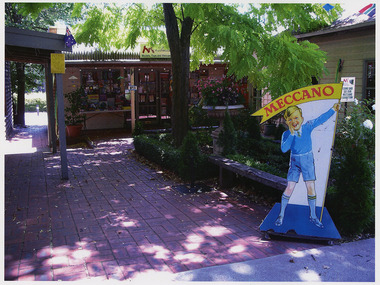 Shows a large advertising sign with a boy holding a flag that says "Meccano". In the background is the shop front for Manical Mechanicals in Marysville in Victoria.