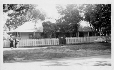 A copy of an old black and white photograph of Carlisle Guesthouse in Marysville. Shows an old weatherboard building with a verandah running all the way around. Shows a wooden picket fence running along the front of the property. Shows a lady and a man standing in front of the picket fence.