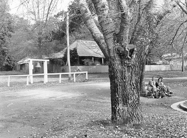 A black and white photograph of the Carlisle Guesthouse in Marysville in 1952. Shows an old building with a verandah running along the front of the building. The property is fronted by a wooden picket fence. In the foreground across the road from the guesthouse, is a group of one man and two ladies sitting on a wooden bench seat.